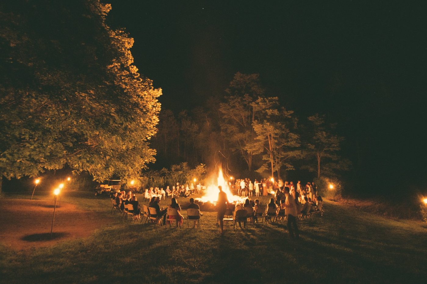 Menschen sitzen nachts im Kreis um ein großes Lagerfeuer unter Bäumen, beleuchtet von Fackeln
