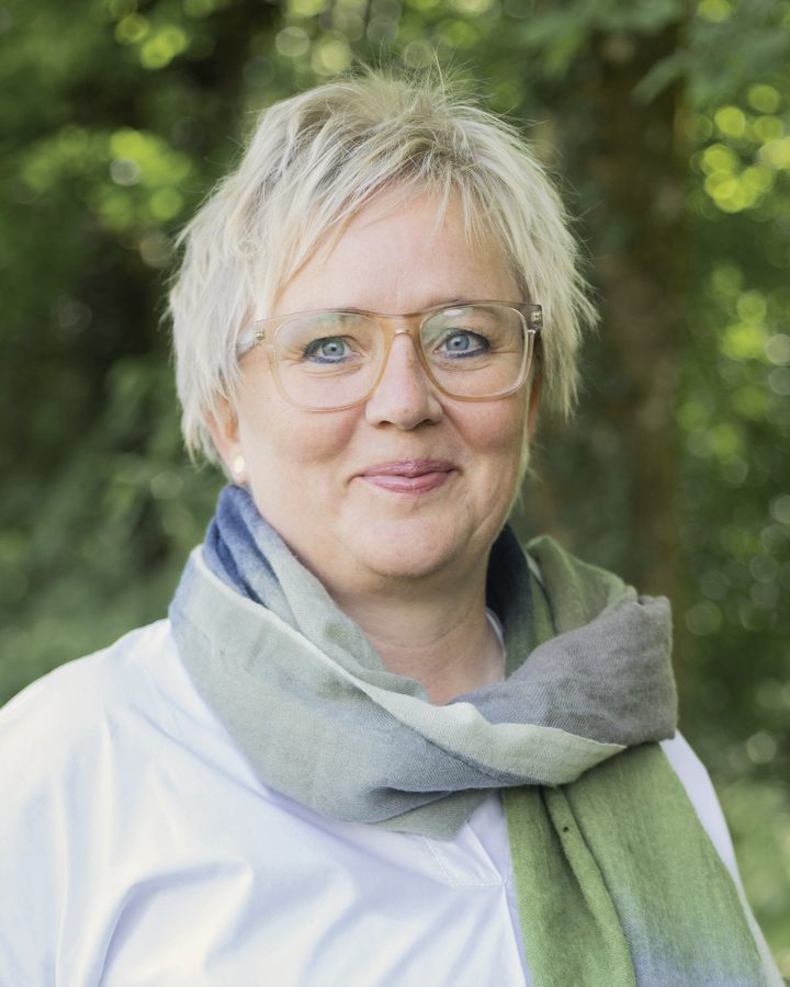 Porträt von Silke Fischer Petersohn: kurze blonde Haare, Brille, weiße Bluse und grüner Schal, im Hintergrund Laub